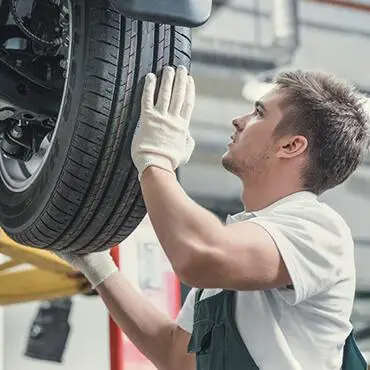 A mechanic wearing gloves inspects a car tire on a lifted vehicle in a garage. He is looking up while examining the tires surface, with various equipment visible in the background.