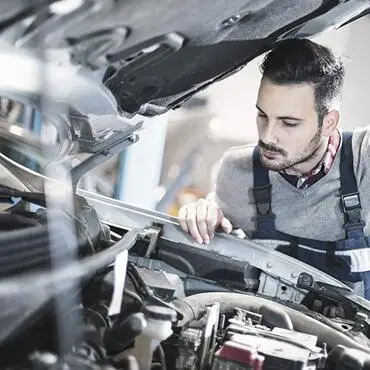 A mechanic in overalls and a long-sleeve shirt inspects the engine of a car. The vehicles hood is open, revealing various engine components. The mechanic appears focused and is looking closely at the engine parts.