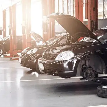 Image of a car repair shop with several vehicles in a row. Each car has its hood open for maintenance. One vehicle in the foreground is elevated with its front wheel removed, exposing the brake disc. Bright lighting streams in from the windows.