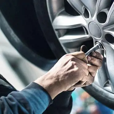 A person using a tool to work on a silver car wheel. Hands are gripping a socket wrench, tightening or loosening lug nuts. The background is blurred, emphasizing the focused activity on the wheel.