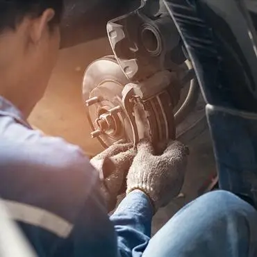 The Mufflerman A mechanic in blue overalls and gloves works on a cars brake system, focusing on the disc and caliper. The scene is lit with warm light, highlighting the details of the vehicles wheel assembly.