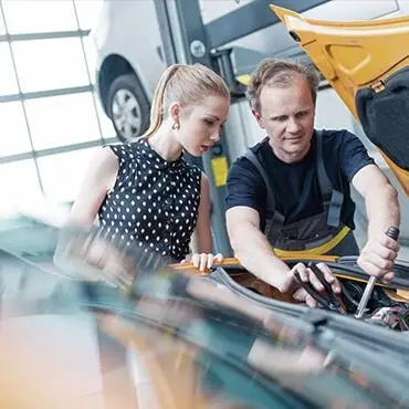 A woman and a mechanic examine the engine of a yellow car in a garage. The mechanic, wearing overalls, works on the engine while the woman in a polka dot dress looks on attentively. A car is visible on a lift in the background.