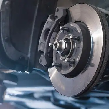 Close-up of a cars brake assembly, showing a shiny metal brake disc mounted on a wheel hub, with a brake caliper positioned above. The background is blurred, highlighting the mechanical components.