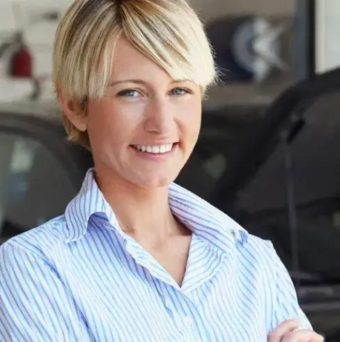 A woman with short blonde hair and wearing a blue and white striped shirt smiles at the camera. She stands in front of a blurred car in a garage.