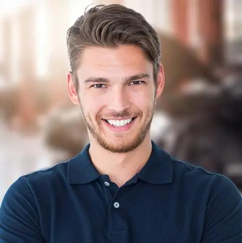 Smiling person with short, light brown hair and facial hair, wearing a navy blue polo shirt. The background is blurred and appears to be an urban or city setting with buildings.