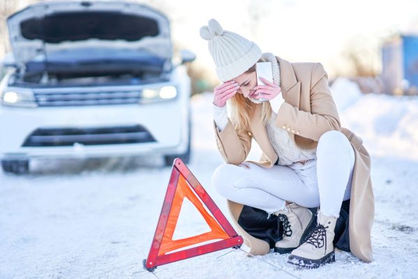 The Mufflerman, Mechanic Near Me A woman kneels on a snowy road, phone in hand, looking distressed with one hand on her forehead—a reminder of the importance of winter vehicle maintenance tips. Nearby, a red triangular warning sign sits on the ground with a white car parked behind her, its hood open.