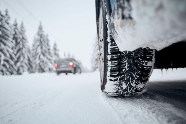 The Mufflerman, Mechanic Near Me Close-up of a car tire on a snowy road, exemplifying Winter Tires 101 essentials, with snow-covered trees in the background. Another vehicle is driving away in the distance. The snow creates a cold, wintry atmosphere.